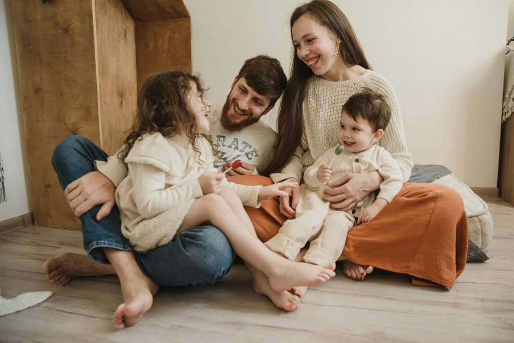 Eine lächelnde vierköpfige Familie sitzt auf dem Wohnzimmerboden und genießt die gemeinsame Zeit.